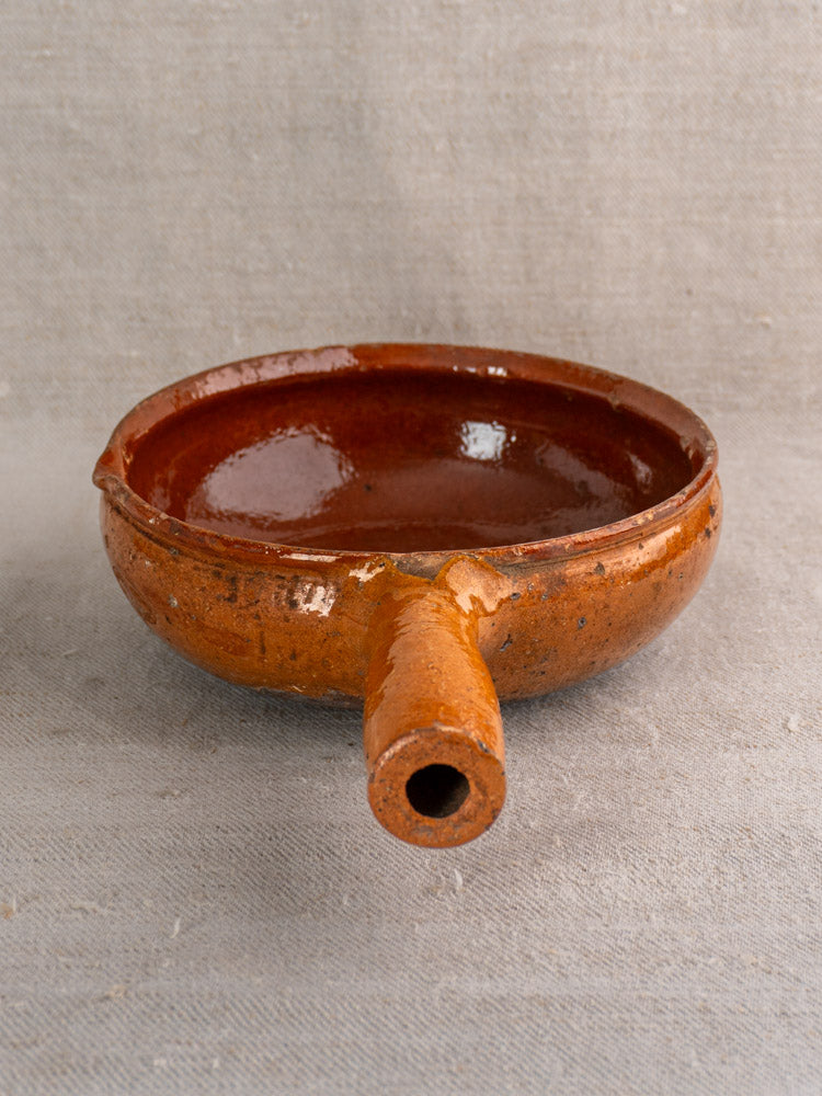 Antique French Vallauris poêlon, brown glazed earthenware with side spout, resting on textured beige fabric.