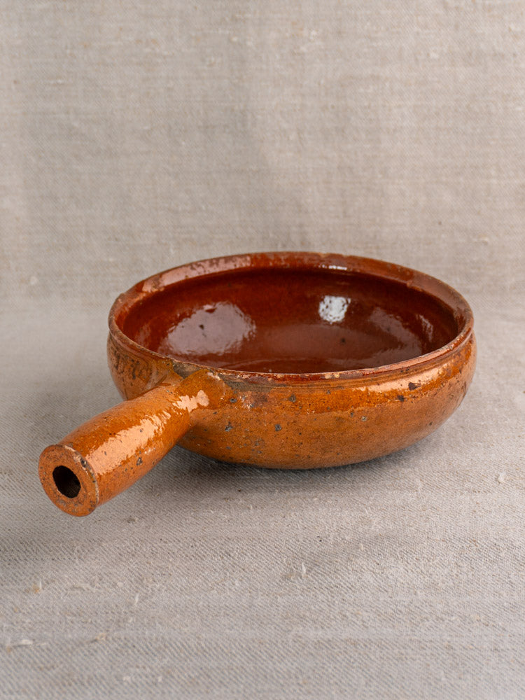 Antique French Vallauris glazed earthenware poêlon with short handle on textured beige fabric.