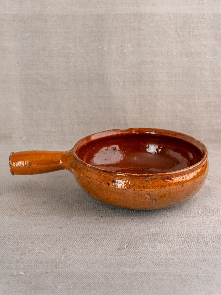 Handmade antique French Vallauris earthenware poêlon with glossy brown glaze on textured beige fabric.