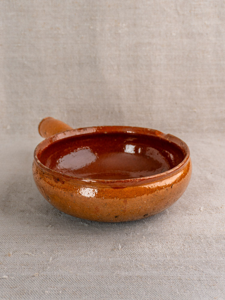 Antique French Vallauris glazed earthenware poêlon, round and brown with a short handle, on textured beige fabric.