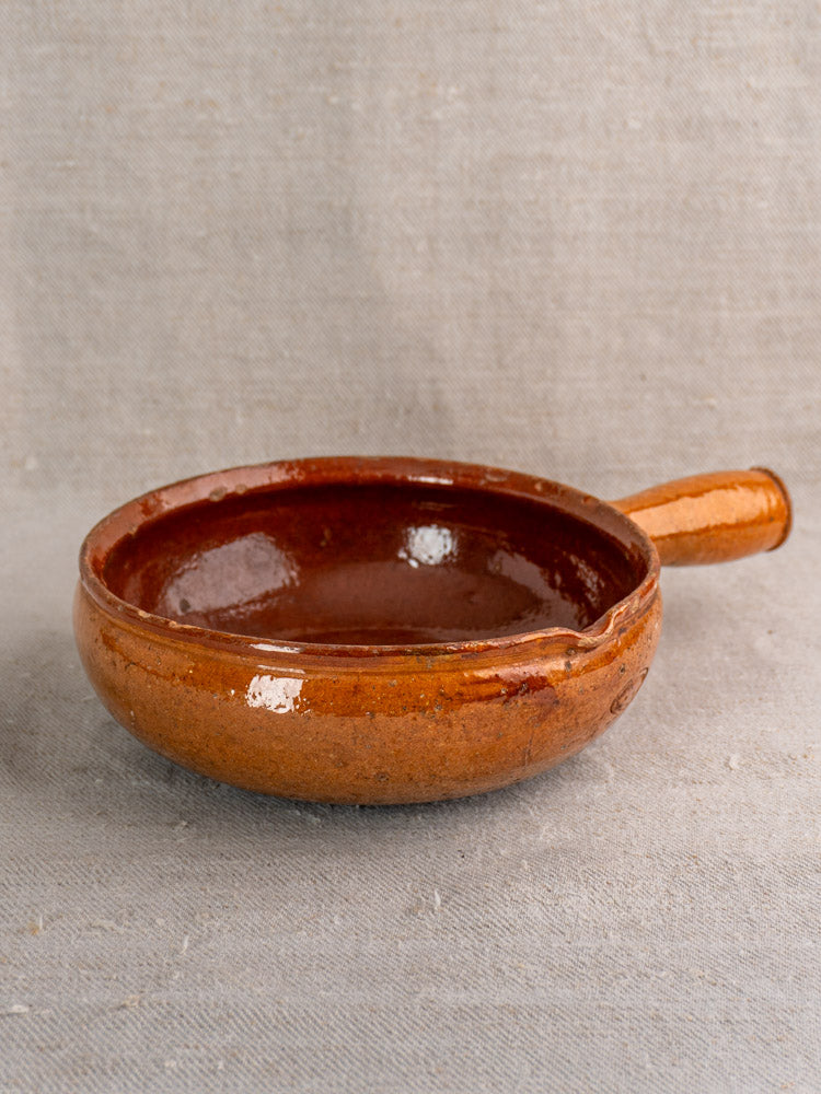 Antique French Vallauris glazed earthenware poêlon with handle and spout on textured gray fabric background.