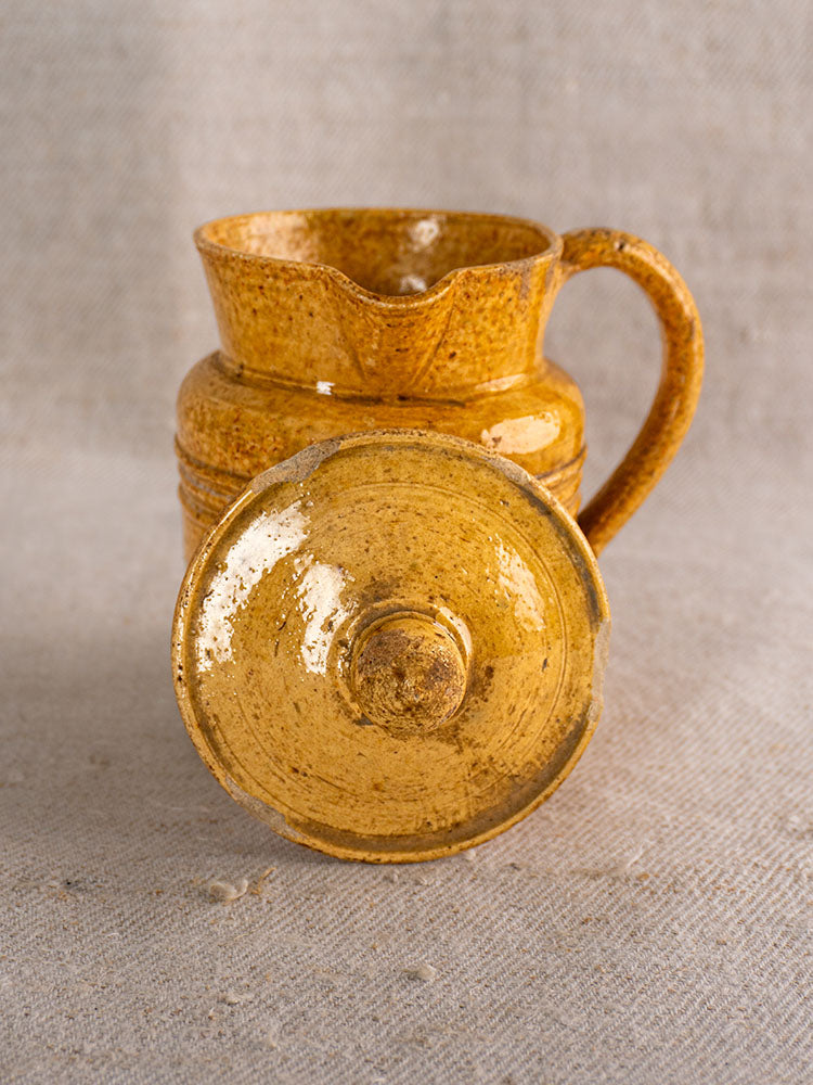 Antique French Pot à Lait with speckled yellow glaze and ornate handle on textured beige fabric.