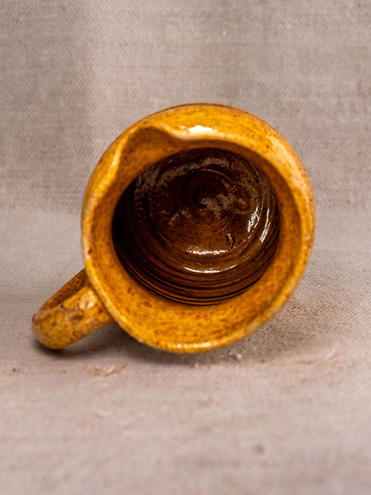 Antique French pot à lait with speckled yellow glaze, lid, and handle, atop coarse beige fabric.