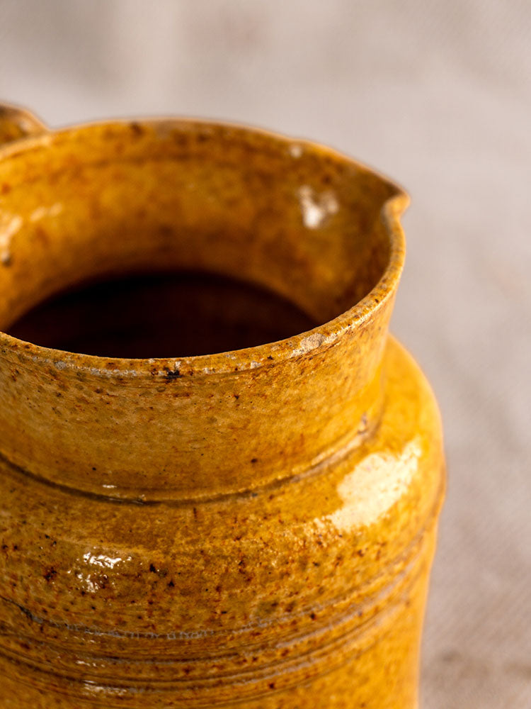 Antique French Pot à Lait with speckled yellow glaze and lid, rim and spout shown, blurred neutral background.