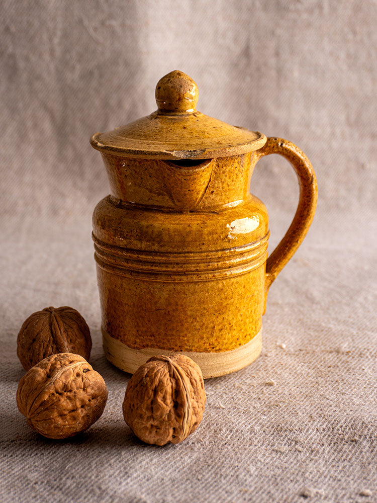 Antique French pot à lait with speckled yellow glaze and lid on textured fabric beside three walnuts.
