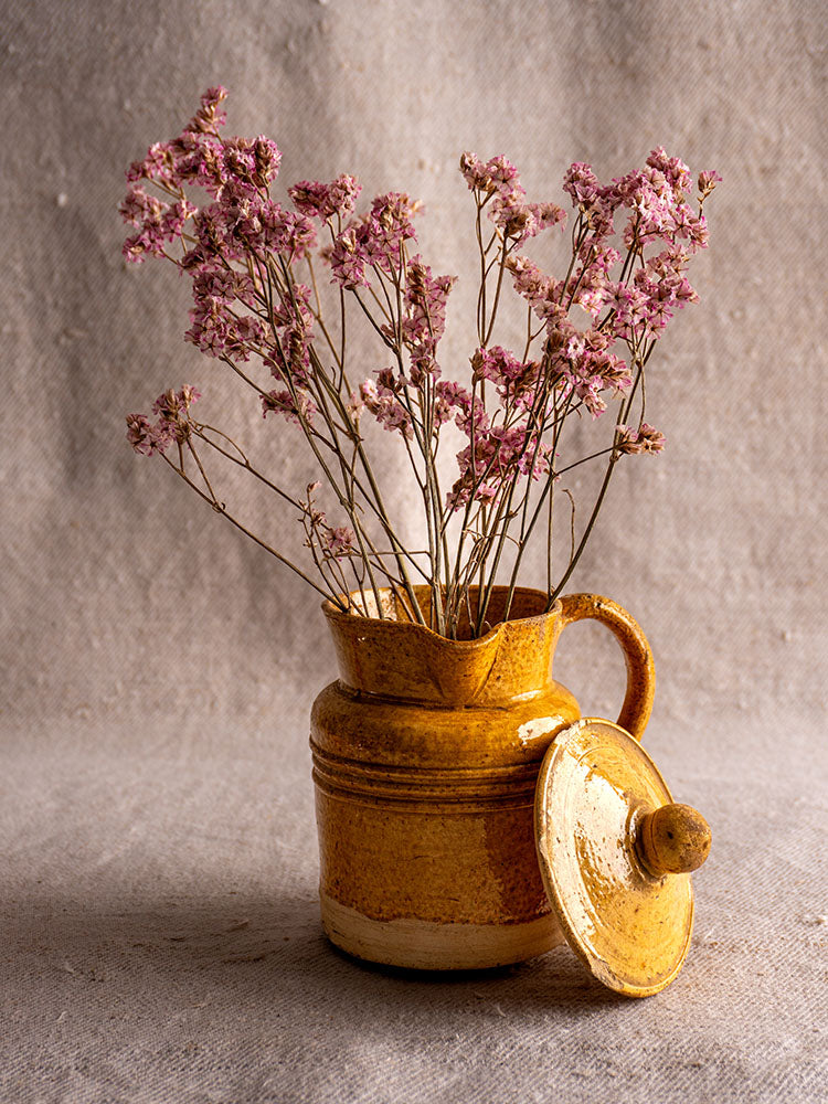 Antique French pot à lait with speckled yellow glaze and lid, filled with pink dried flowers, on neutral background.