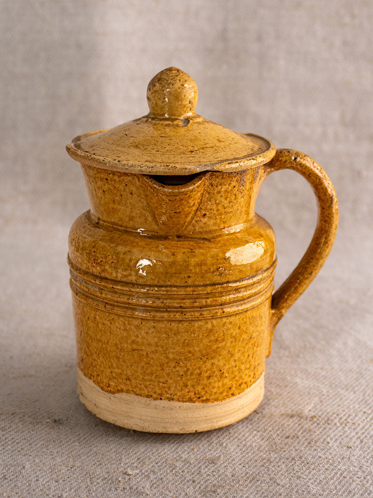 Antique French pot à lait with yellow speckled glaze, lid, and handle displayed on textured beige fabric.
