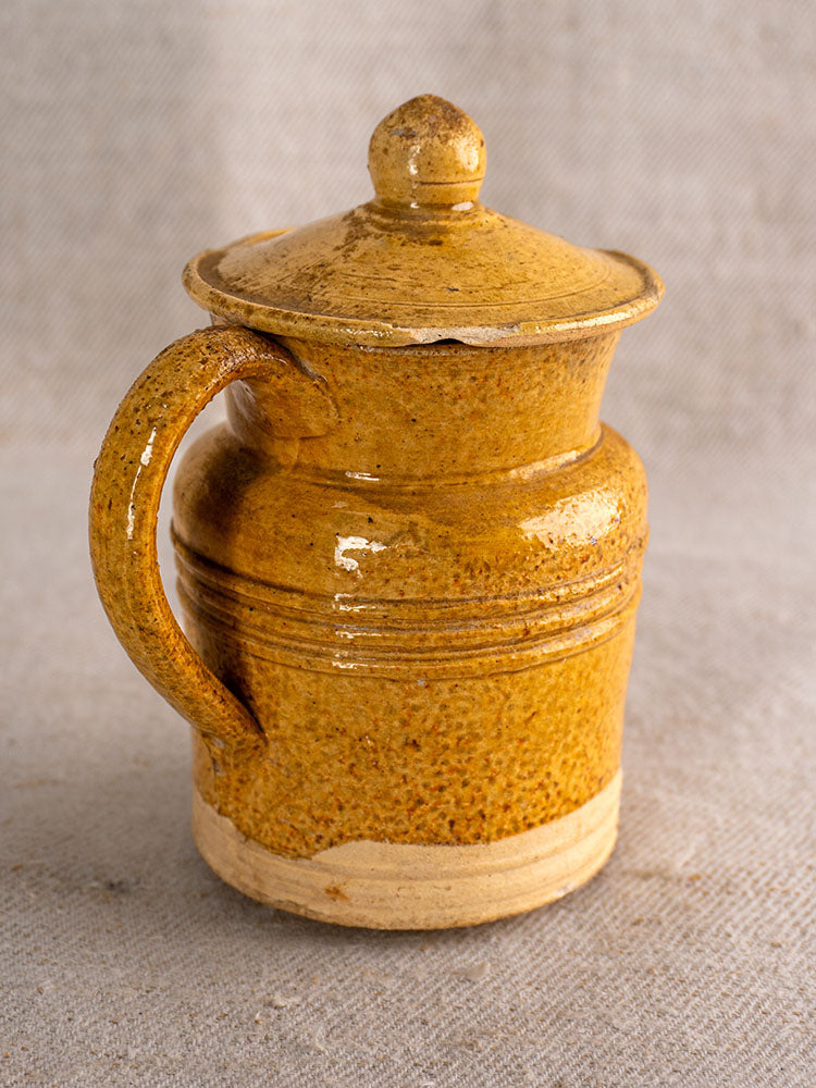 Antique French Pot à Lait with speckled yellow glaze, lid, and handle on a light fabric backdrop.