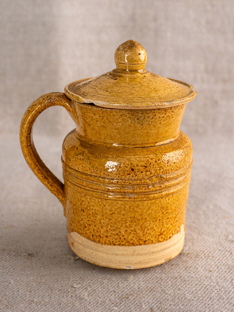 Antique French pot à lait with speckled yellow glaze, grooved detail, lid, and handle on textured fabric background.