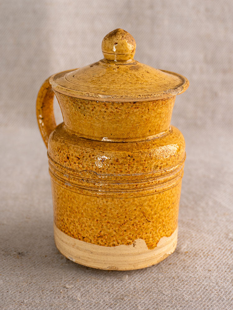 Antique French pot à lait with speckled yellow glaze, lid, and handle on textured gray fabric. Glossy with unglazed base.