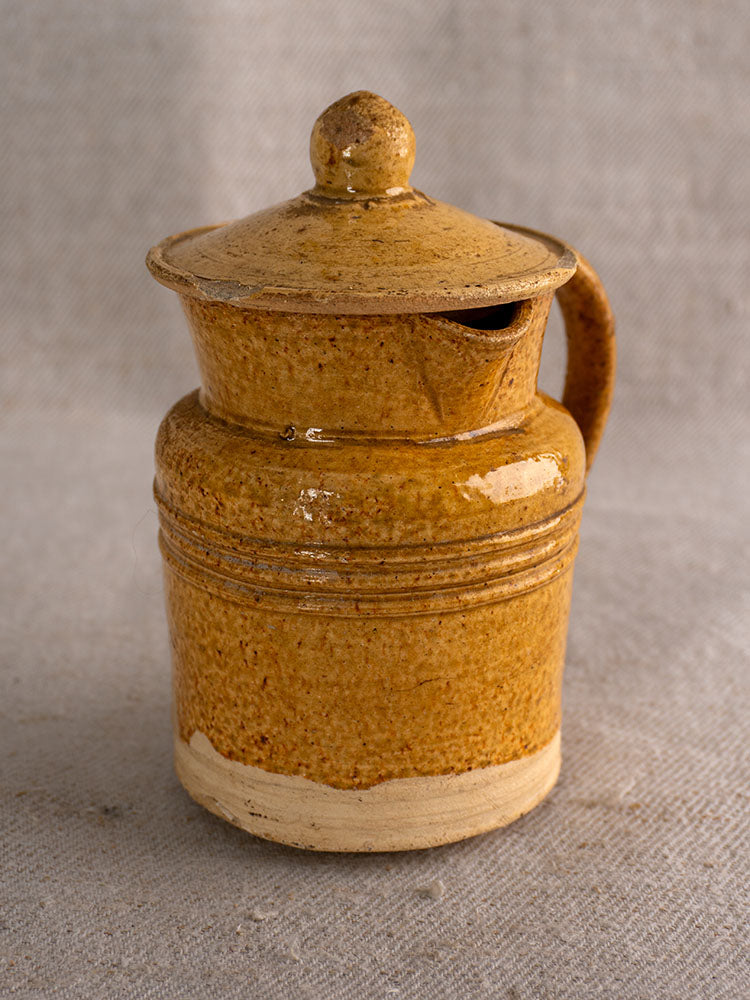 Antique French Pot à Lait with speckled yellow glaze, curved handle, lid, and spout on beige textured fabric.