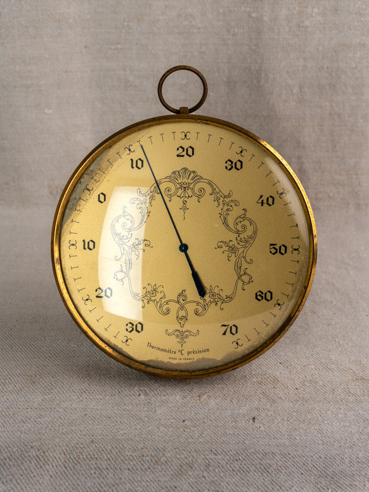 Vintage French gilt wall thermometer with ornate gold dial, Celsius display, on beige fabric background.