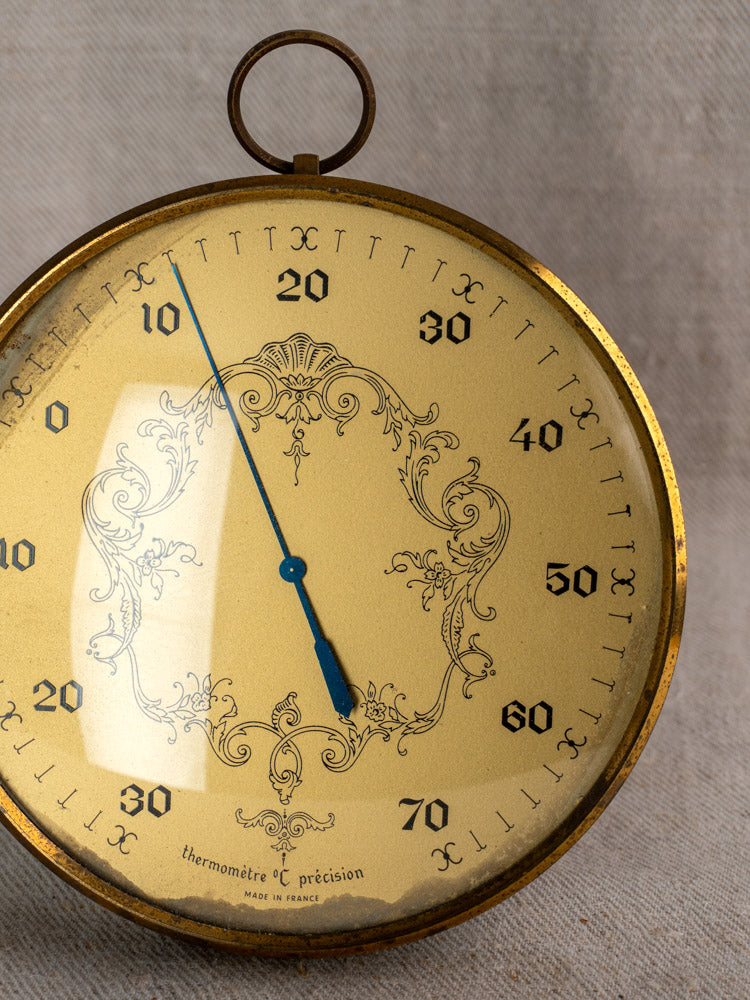 Vintage French gilt wall thermometer with ornate detailing, blue needle at 18°C, on grey fabric backdrop.