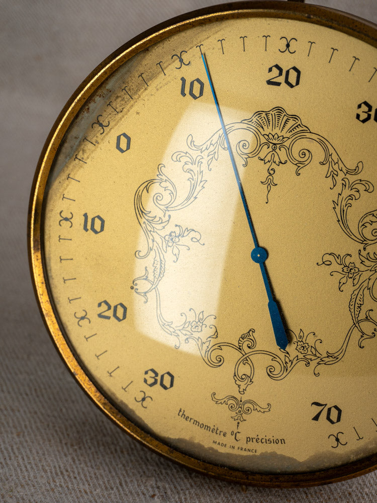 Vintage French gilt wall thermometer with ornate gold-tone frame and blue needle, beige fabric background.