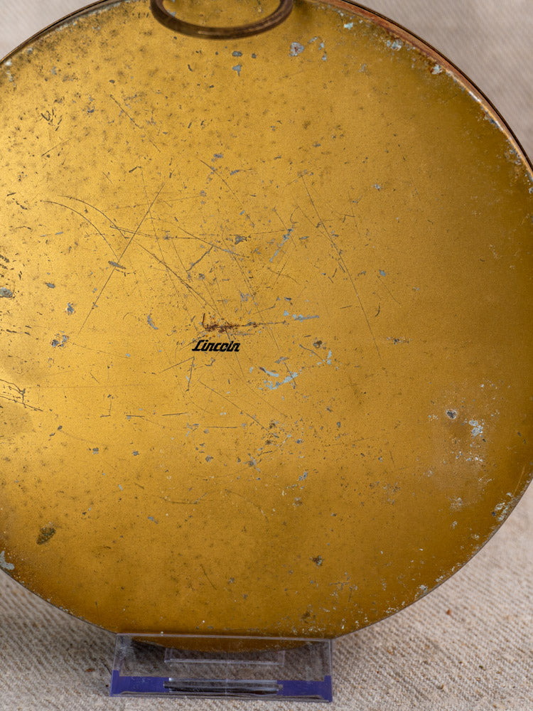 Vintage French Lincoln gilt wall thermometer with scratched gold-tone metal face and central black script, on fabric surface.