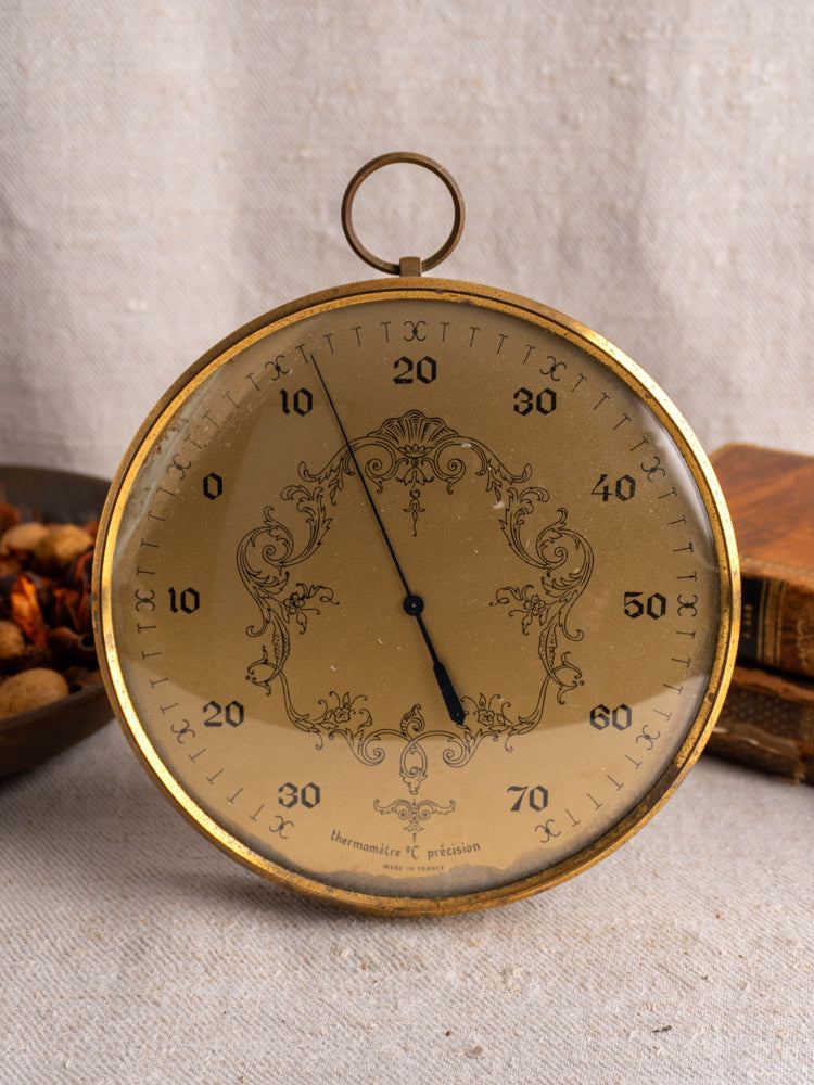 Vintage French Lincoln thermometer with gilt wall dial, displayed on beige fabric near wooden bowl and old book.