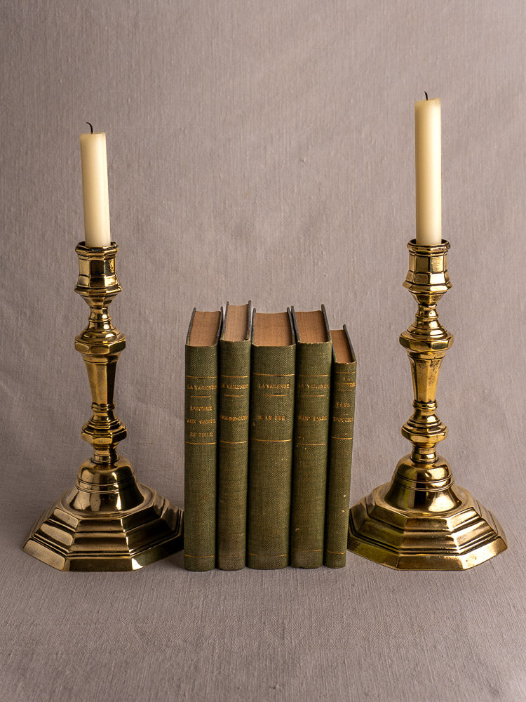 Pair of Régence period antique French brass candlesticks with white candles, flanking green hardcover books on beige fabric.