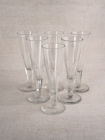 Set of six early 19th century antique French cut crystal champagne flutes on light beige textured surface with fabric backdrop.