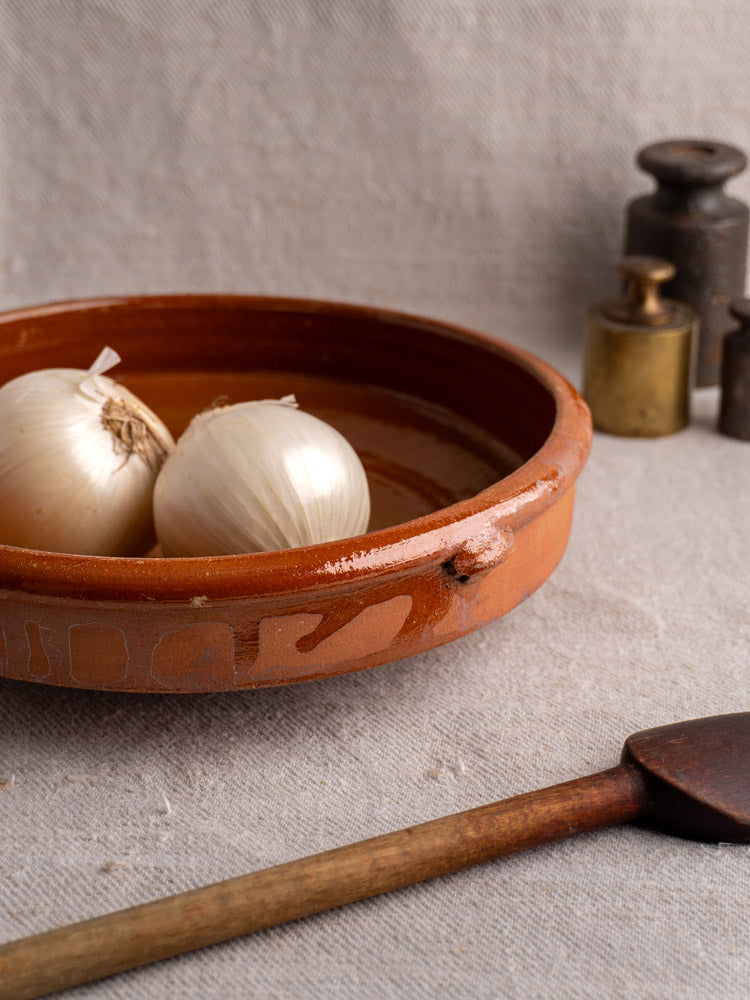 Antique French Tian glazed terracotta baking dish with onions, vintage spatula, and metal weights on fabric.
