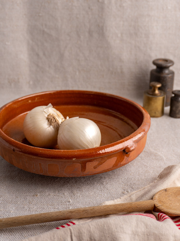 Antique French Provençal glazed terracotta Tian baking dish with white onions on beige fabric, wooden spoon and metal weights nearby.