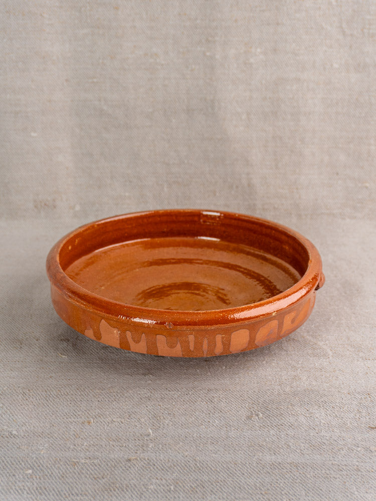 Antique French terracotta tian with glossy brown glaze and rim notch on textured beige fabric background.