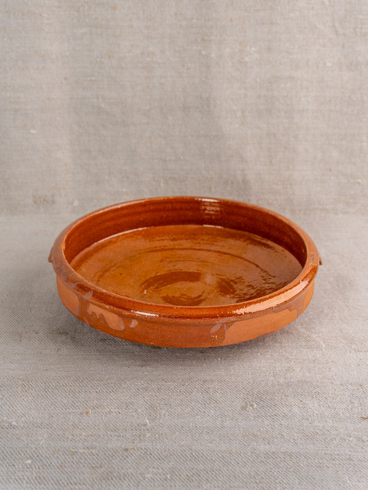 Antique French glazed terracotta Tian Provençal baking dish, round and shallow, glossy brown, on light gray textured fabric.
