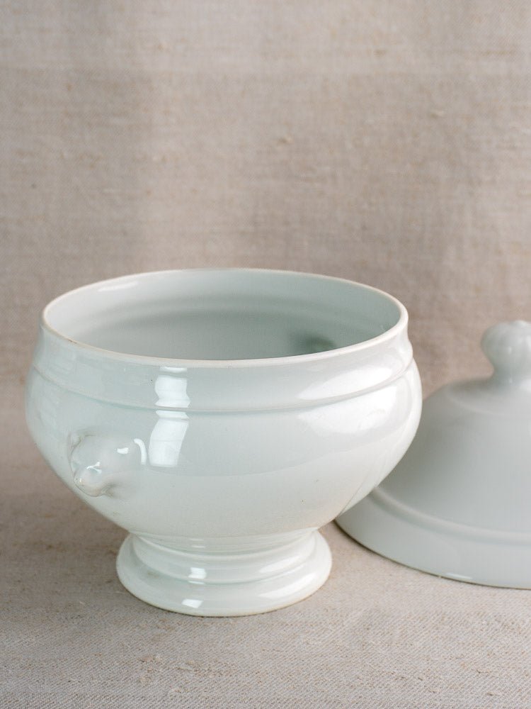 Vintage French white ceramic soup tureen with lid, shown with twelve lion head bowls on beige fabric.