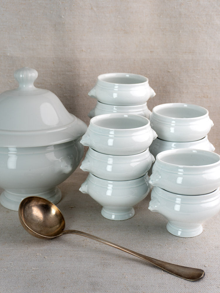 Vintage French CNP soup tureen with twelve lion head bowls and metal ladle on neutral fabric.