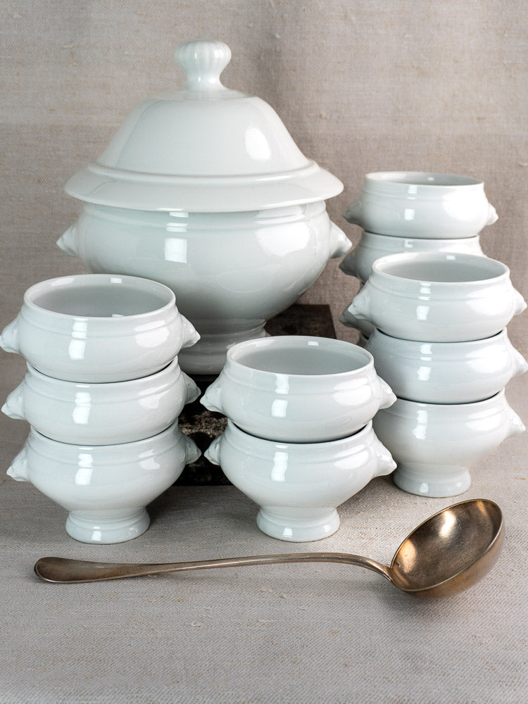 Vintage French CNP soup tureen with twelve lion head bowls and metal ladle on neutral fabric.