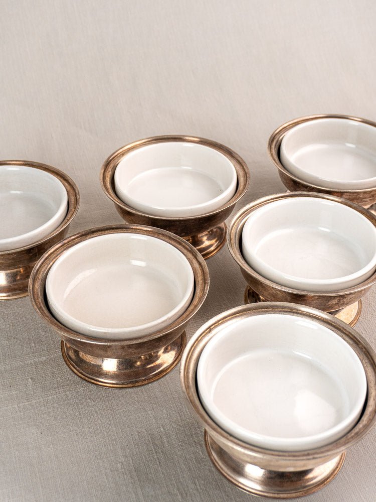 Five vintage Sambonet silver-plated coupes with porcelain inserts on a light fabric surface.
