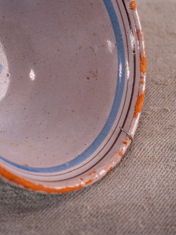 19th century French Cul Noir faience bowl with blue floral motifs, chipped orange rim, metal staple repair on gray fabric.