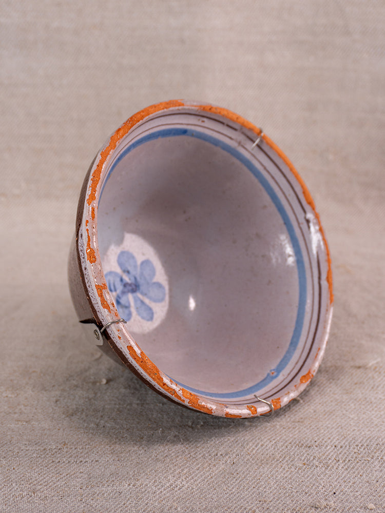 Antique French Cul Noir faience bowl with blue floral design, chips, metal staple repairs, and black glazed base on beige surface.