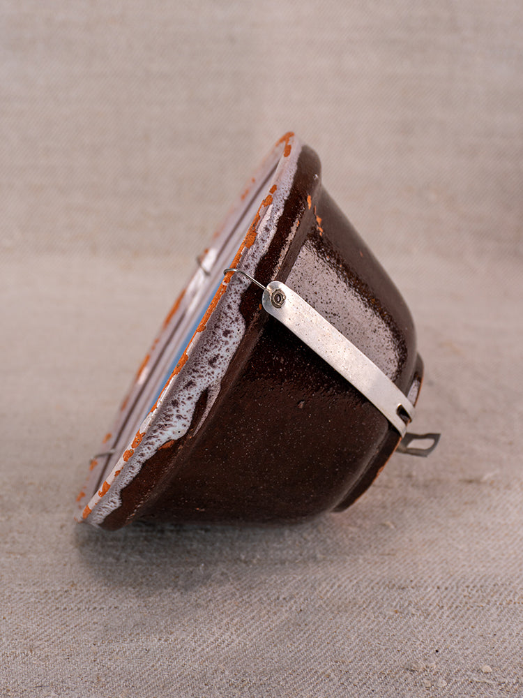 Antique French Cul Noir faience bowl with blue floral motifs, metal clasp and handle, on textured beige surface.