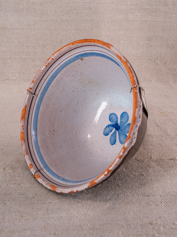 Antique French cul noir faience bowl with blue floral design, staple repair, and chipped edges on beige textured surface.