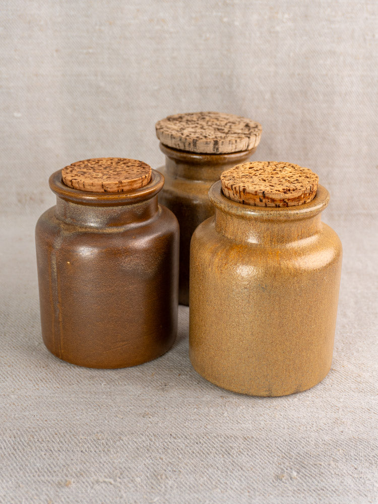 Set of three mid-20th century French stoneware mustard pots with cork lids in earthy brown tones on beige fabric.