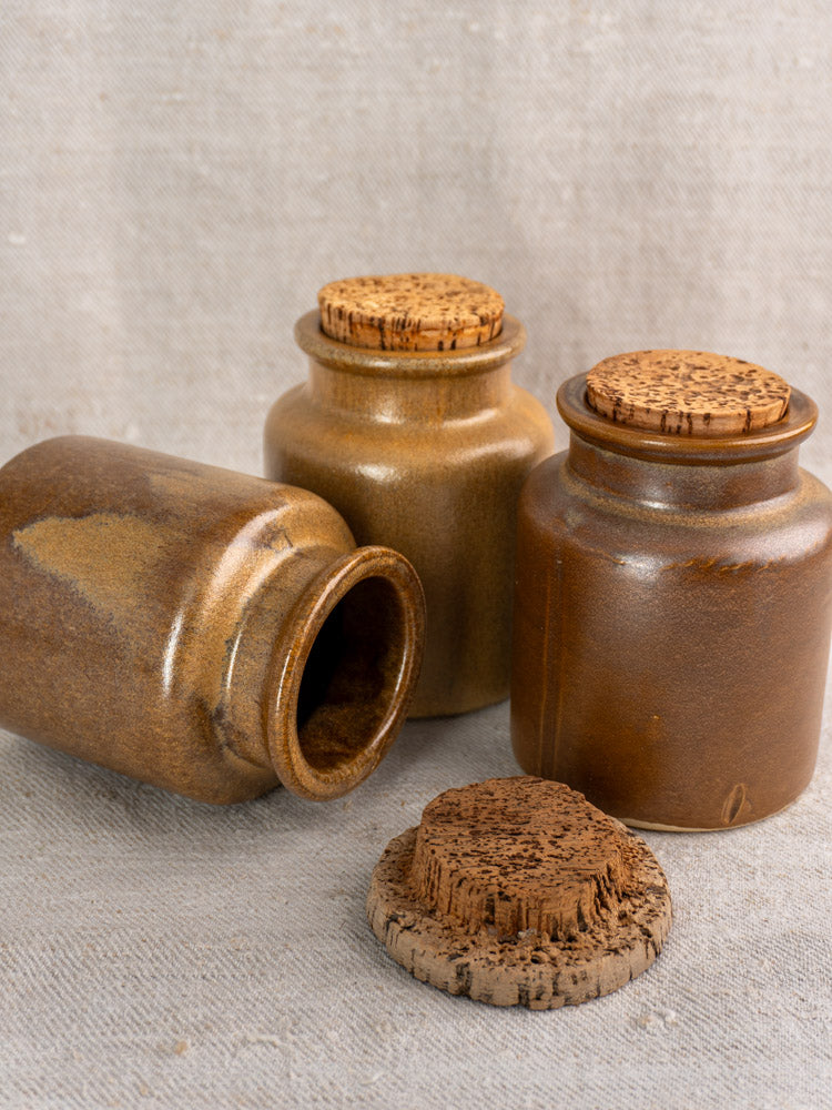 Set of three mid-century French mustard pots, brown stoneware with cork lids, displayed on beige fabric.