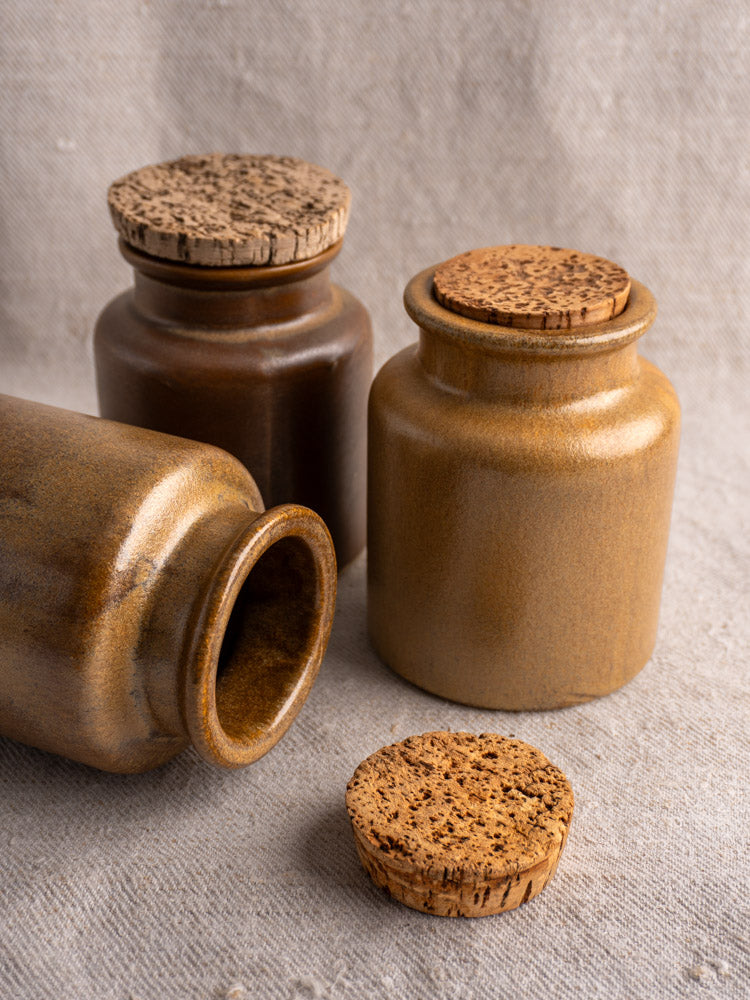 Set of three antique French stoneware mustard pots with cork lids on textured beige fabric, two upright, one lying down.