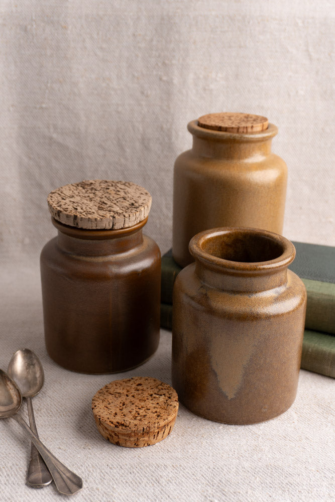 Set of three mid-20th century French stoneware mustard pots with cork lids, displayed on beige fabric with books and metal spoons.