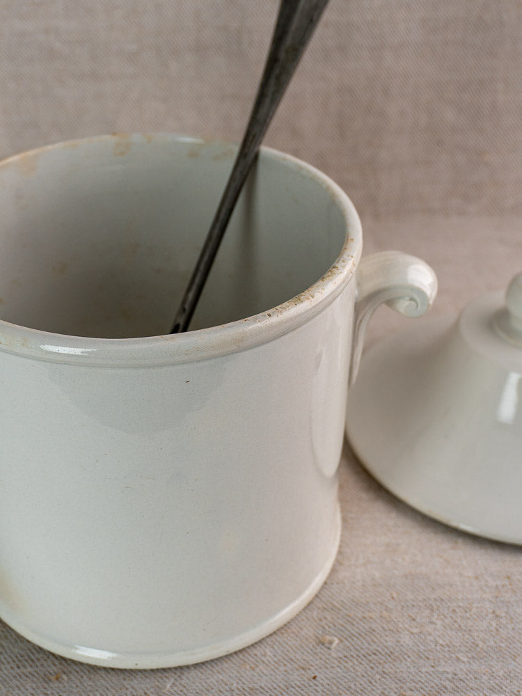 Antique French white faience soup tureen with lid off and metal spoon inside, on neutral surface.