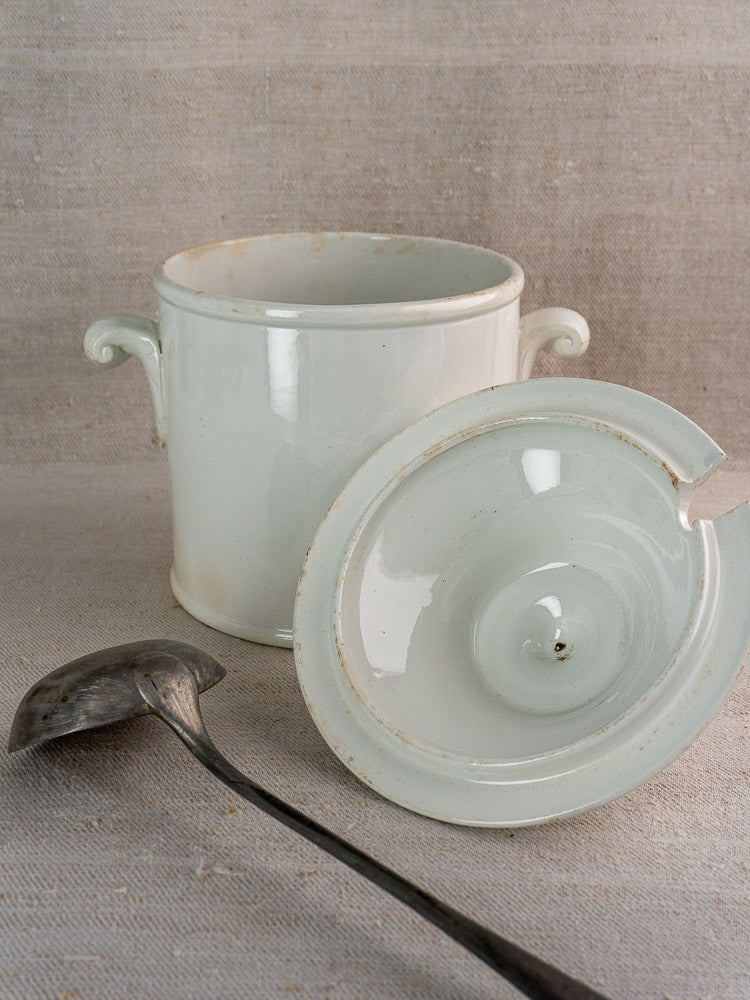 Antique French Lunéville white faience soup tureen with lid and vintage metal ladle on beige fabric.
