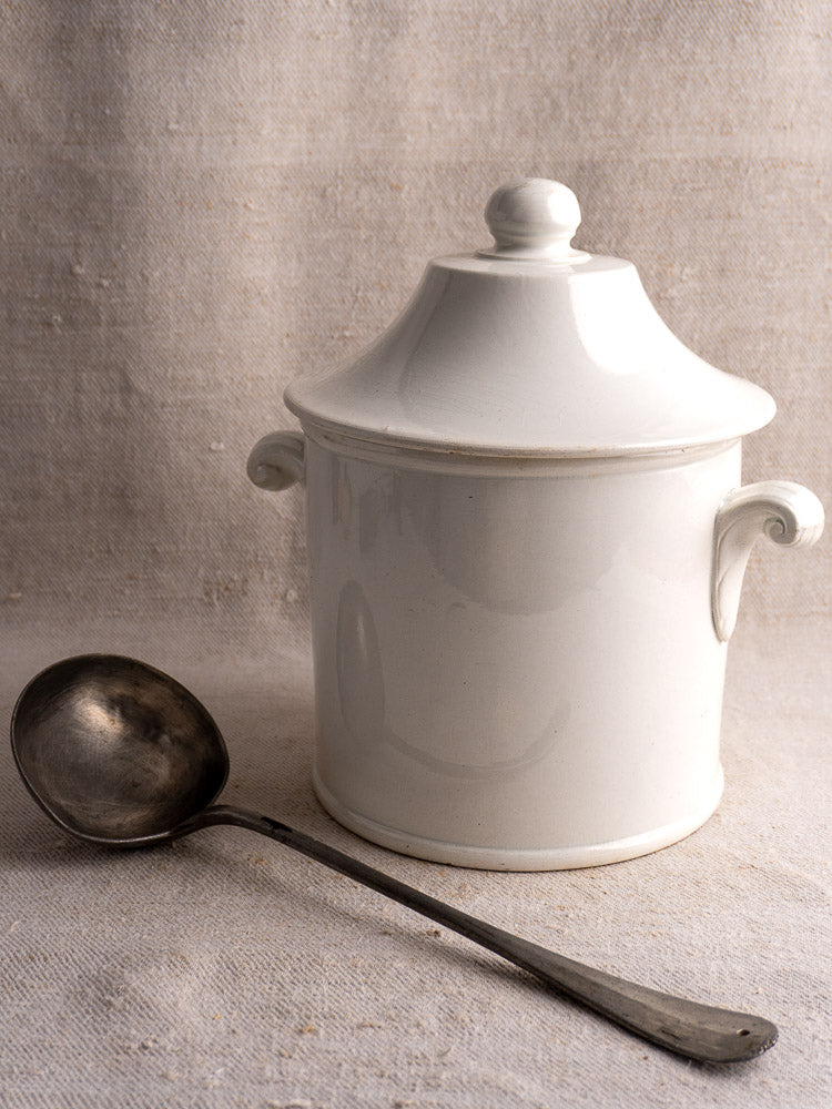Antique French Lunéville white faience soup tureen with matching lid and elegant handles, beside metal ladle on neutral fabric.