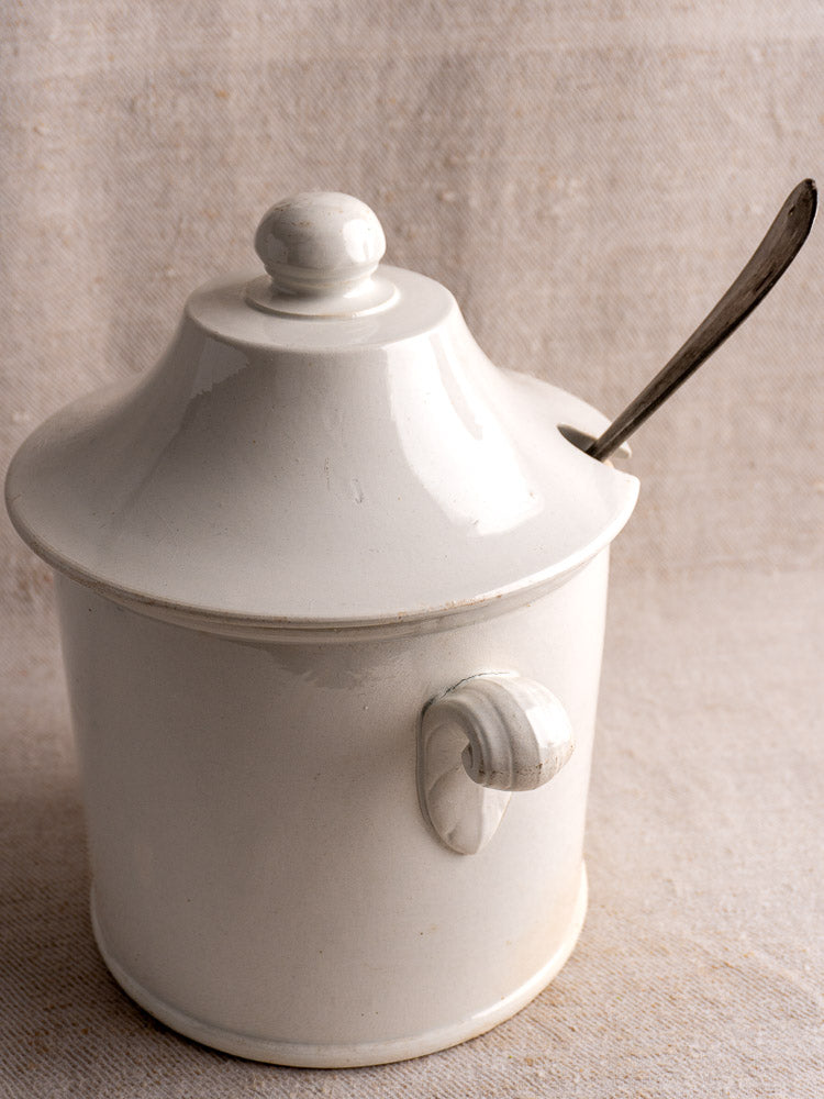 Antique French Lunéville white faience soup tureen with lid, handle, and metal spoon on neutral fabric.