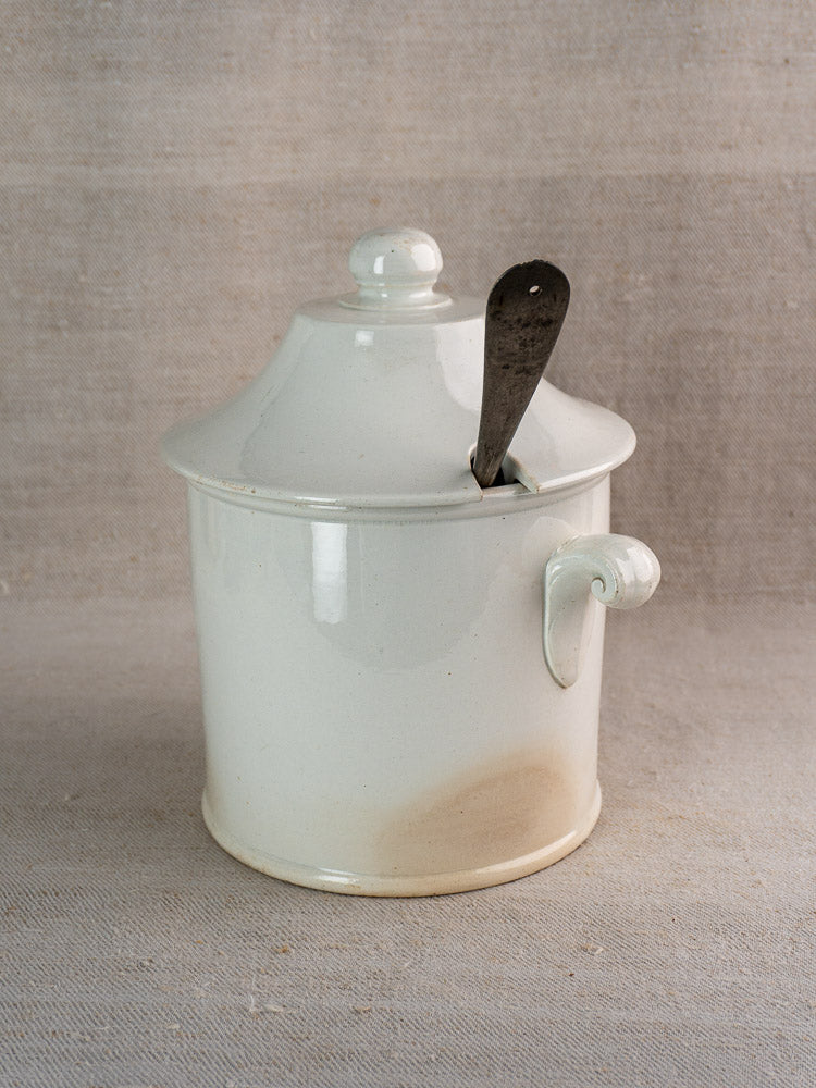 Antique French Lunéville white faience soup tureen with lid and handle, metal spoon inside, on neutral fabric.