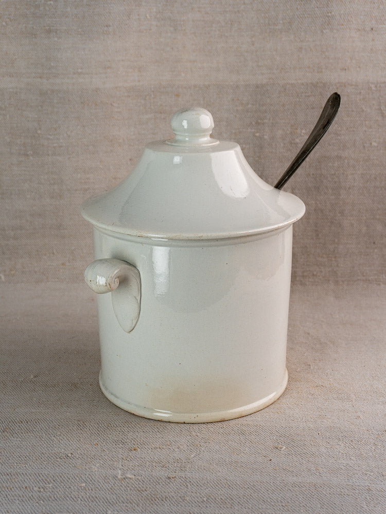 Antique French Lunéville white faience soup tureen with lid and side handle, on neutral surface, metal spoon tucked underneath.