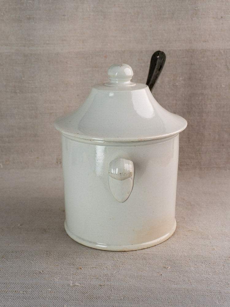 Antique French Lunéville white faience soup tureen with lid and handle, black spoon inside, neutral fabric backdrop.