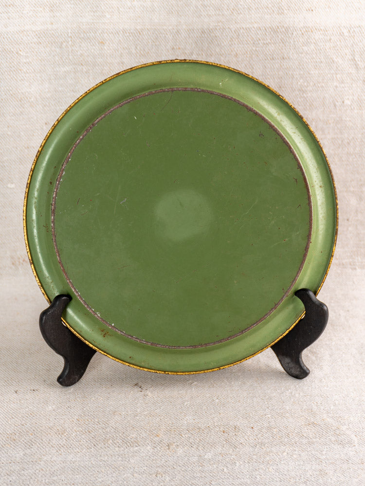 Antique French green painted tôle plate with worn gold rim on black stand, neutral backdrop.