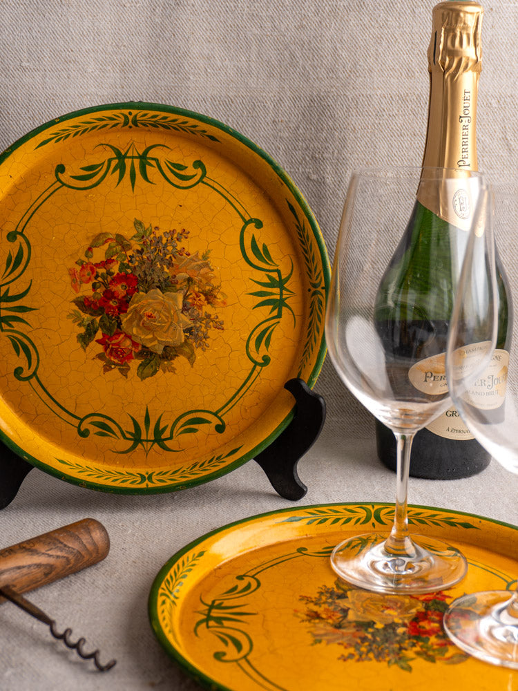 Antique French painted tôle trays with floral motifs, circa 1900, beside champagne and wine glasses.