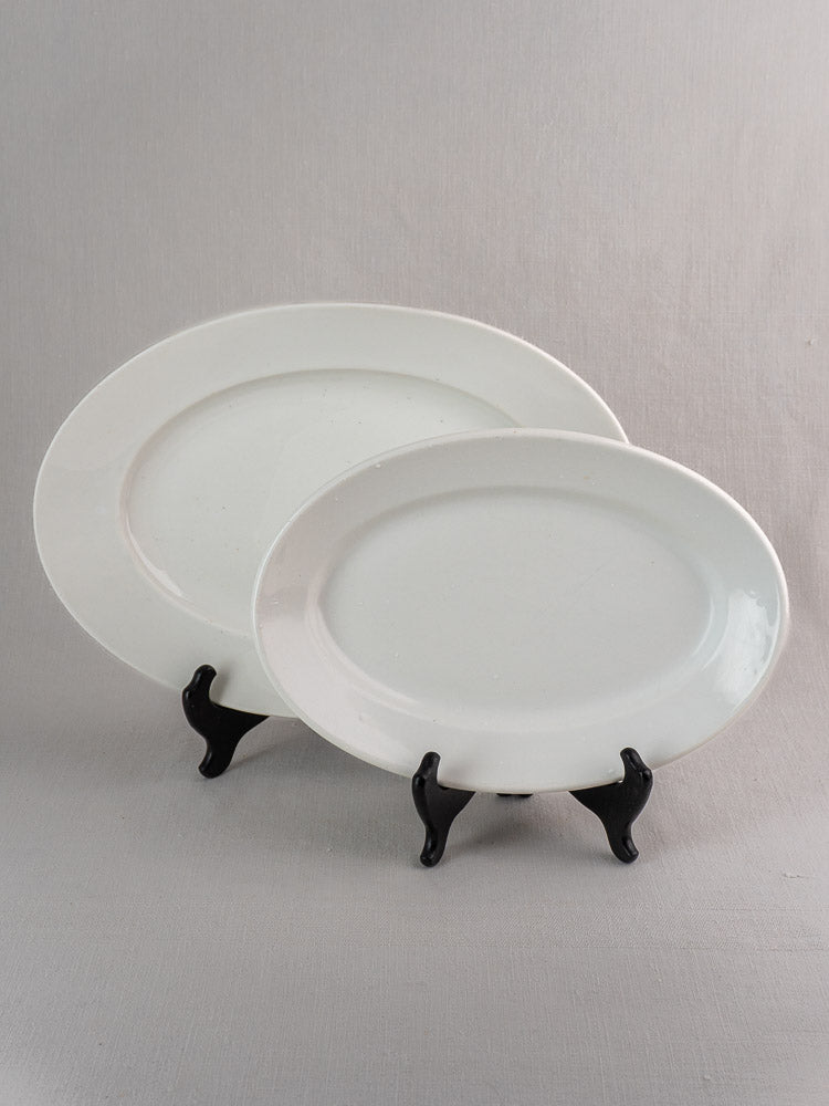 Pair of antique French white ironstone oval platters in graduated sizes on black stands, gray background.