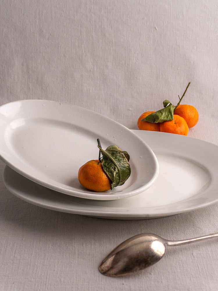 Antique French white ironstone oval platters stacked on grey cloth with clementines and silver spoon.