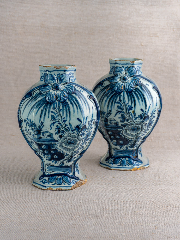 Pair of antique Dutch Delft blue and white vases with floral and landscape motifs on light textured fabric.